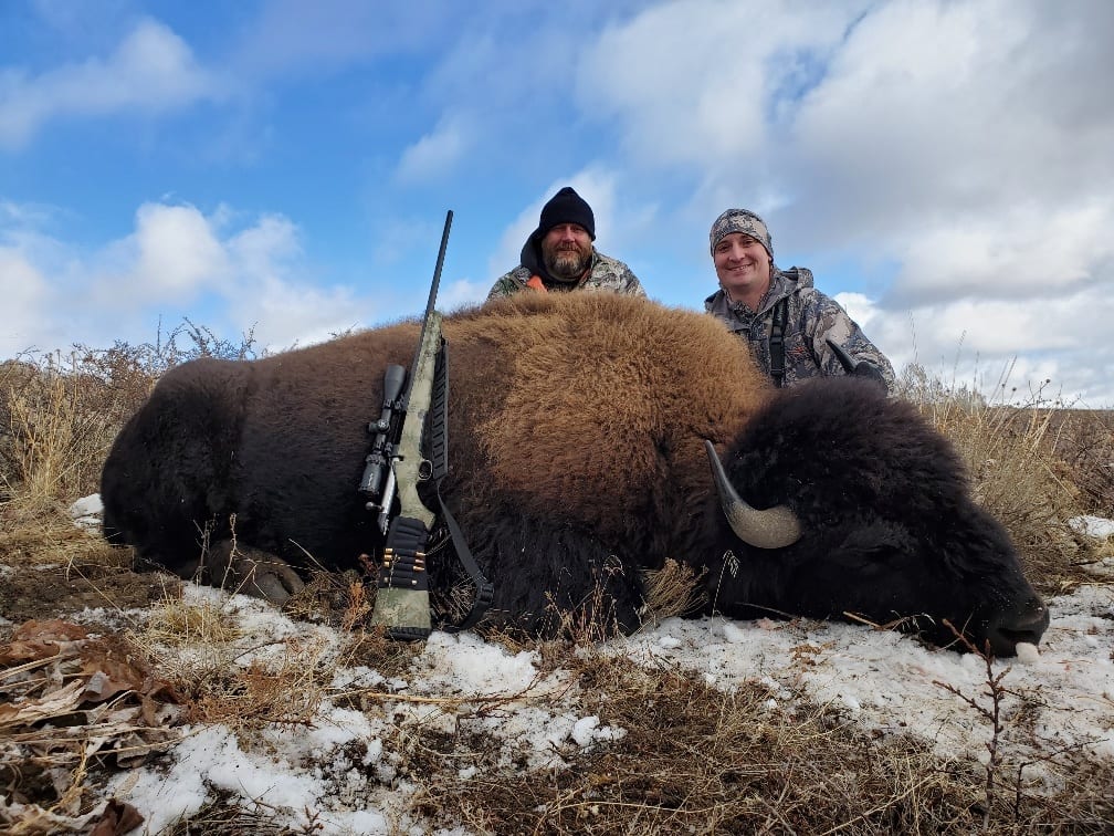 Two hunters pose with a harvested bison in a snowy field.
