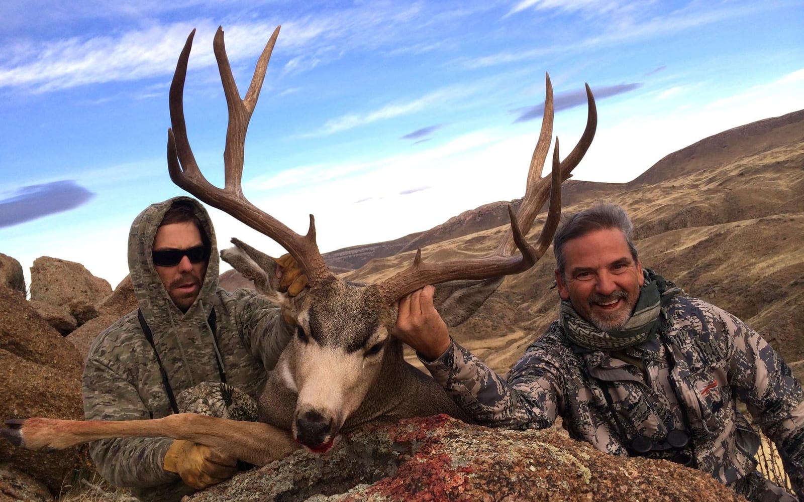 Two hunters pose with a harvested mule deer in a rocky, hilly landscape.