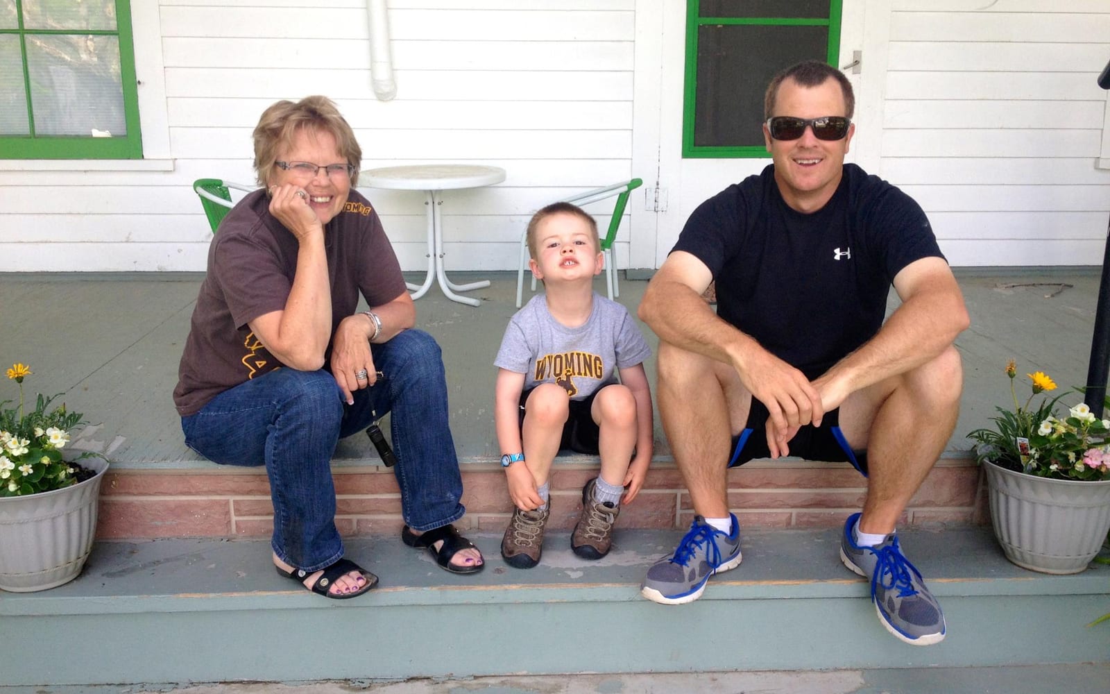 Three people, an older woman, a young boy, and a man, are sitting on a porch with potted plants.