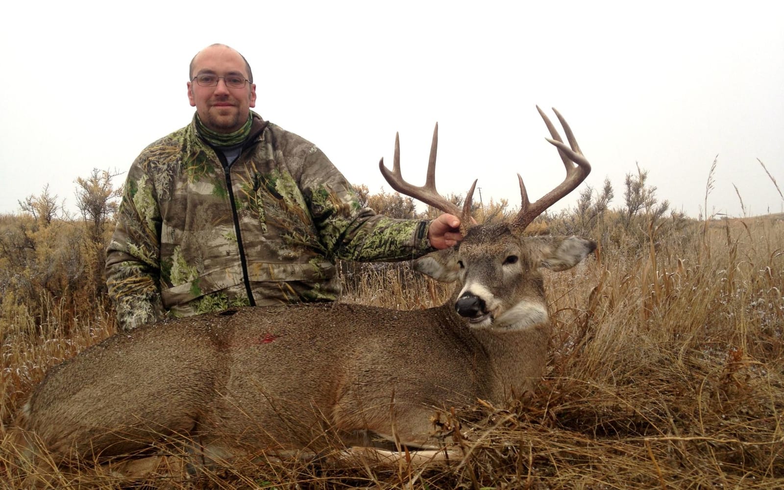A hunter in camouflage poses with a harvested mule deer in a field.
