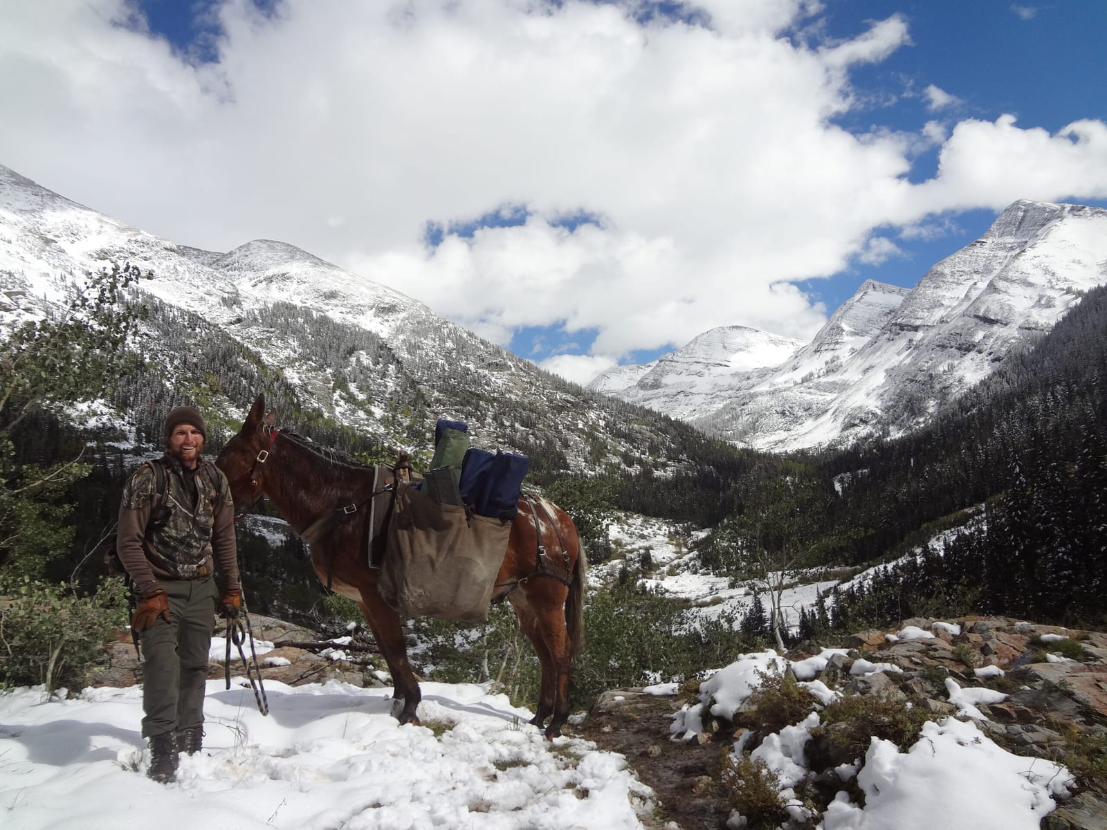 A man in camouflage stands next to a packed mule in a snowy mountain landscape.