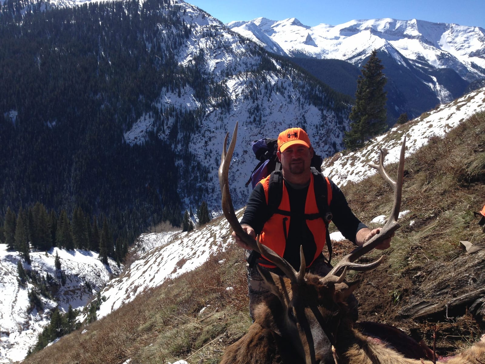 A hunter poses with a harvested elk in a snowy mountain landscape.