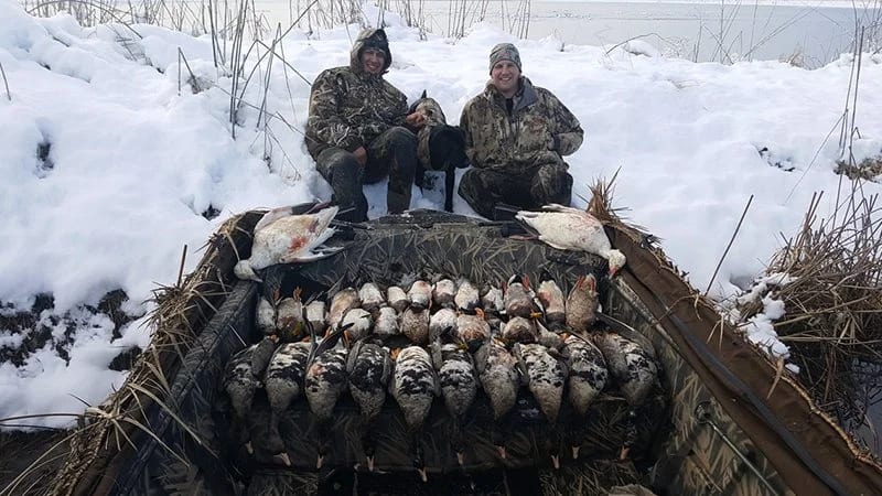 Two hunters in camouflage sit on a snow-covered bank near Lake Shasta, proudly displaying a mixed catch of ducks and fish from their winter outing.