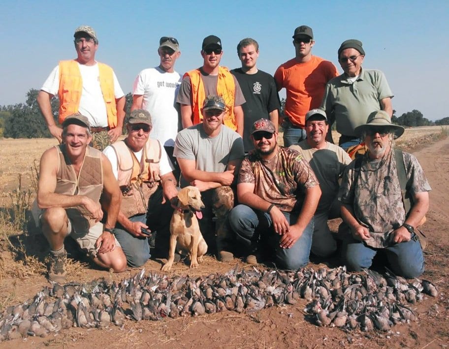 A group of men on a guided hunting trip pose with a large number of birds they harvested, including a dog and a backpack, on a dirt path in a rural area near Klamath Falls, Oregon.