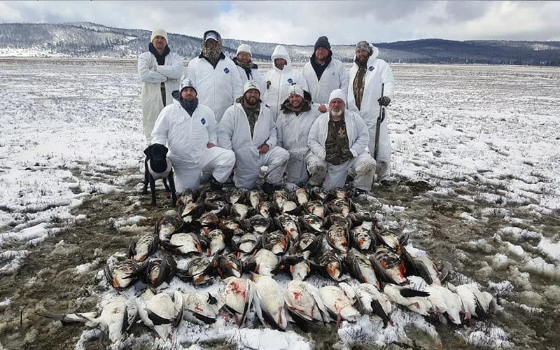 A group of people in white outdoor gear stands on a snowy stretch outside Alturas, California, with a hefty pile of fresh fish at their feet and a dog calmly sitting nearby.