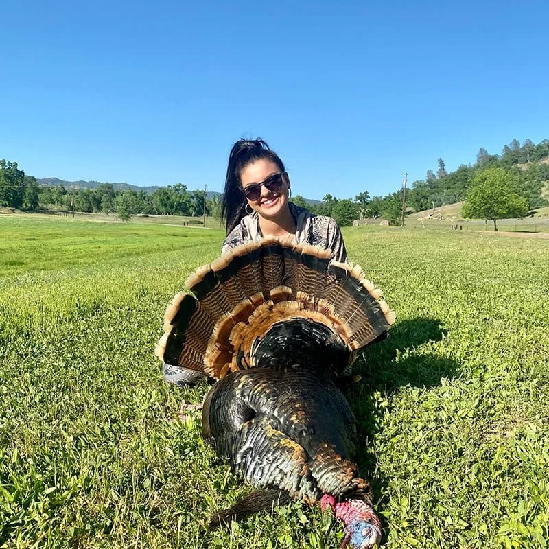 A woman wearing sunglasses kneels next to a large wild turkey in a grassy field framed by the Trinity Alps, the bird’s feathers glinting in the sun.
