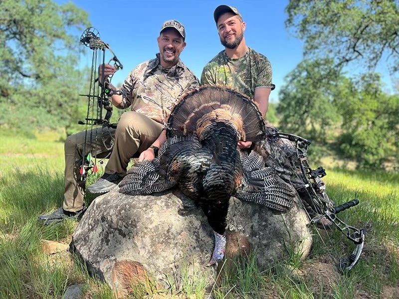Two men in camouflage pose with a large wild turkey they hunted near the outskirts of Yreka, California, their bow and arrow set lying nearby.