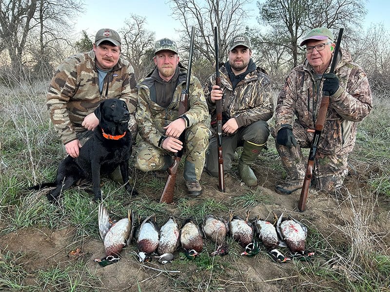 Four men in camouflage and outdoor gear, one holding a rifle, with a black hunting dog sitting beside them. They are kneeling or crouching outdoors in a field with seven ducks laid on the ground in front of them.