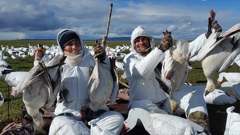 Two hunters in white camouflage suits sit on the open ground near Merrill, Oregon, holding large birds with a field full of snow geese stretching out behind them.