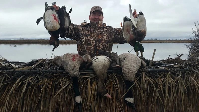 A man in a camouflage jacket and cap holds multiple ducks and geese in front of a marshy waterway near the Scott Valley in Northern California.