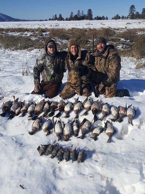Three people in camouflage clothing kneeling on snow in a winter landscape, posing with a large collection of duck decoys or dead ducks laid out in front of them.