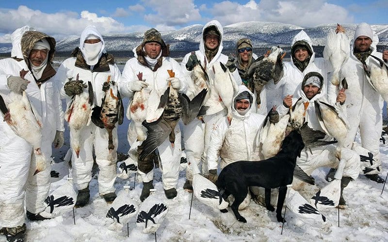 Group of people in white winter gear holding large fish on snowy ground with mountains in the background.