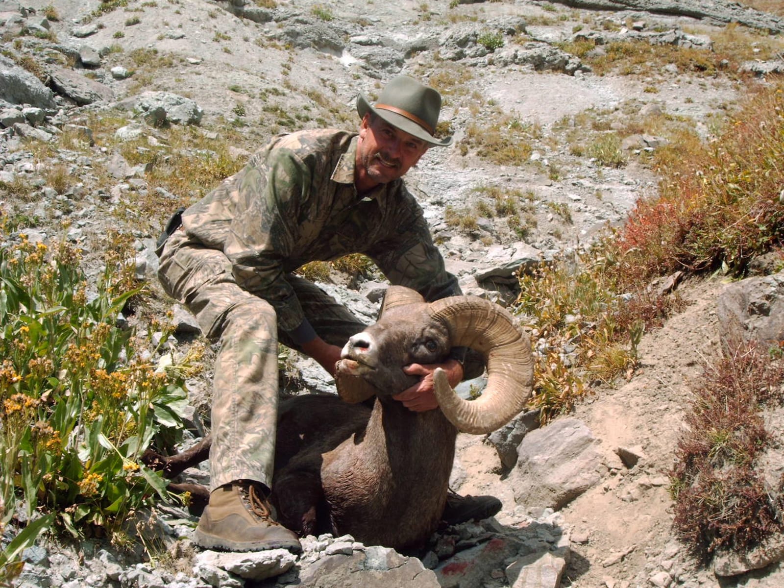 Hunter in camouflage holding a bighorn ram with large curved horns in rocky terrain.