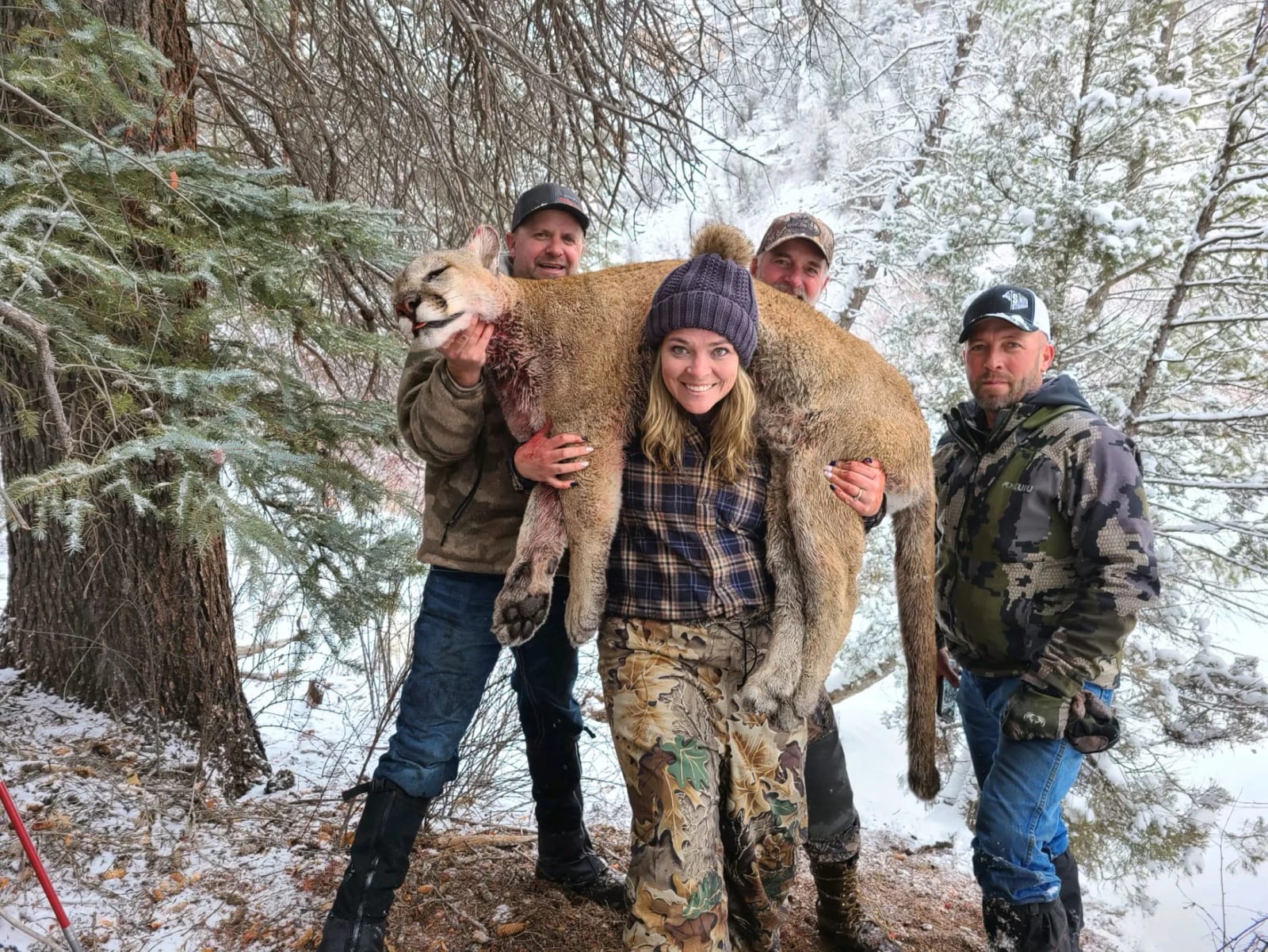 Four people in winter gear holding a dead mountain lion in a snowy forest setting.