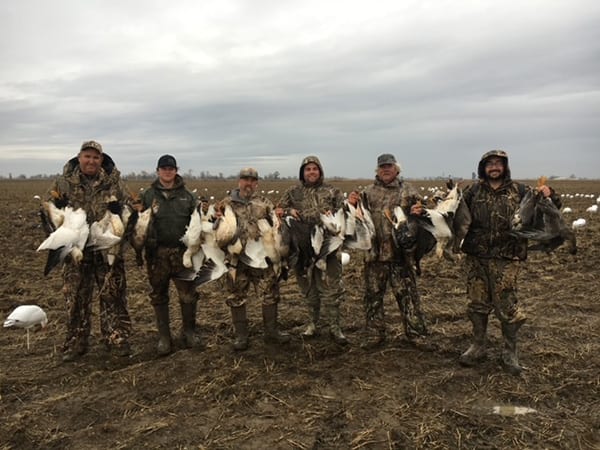 Six hunters in camouflage pose in a muddy field, each holding several harvested snow geese and other geese.