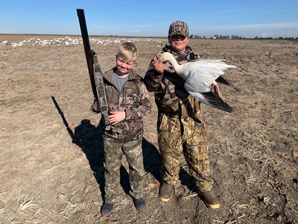 Two young hunters pose with a harvested snow goose in a field.