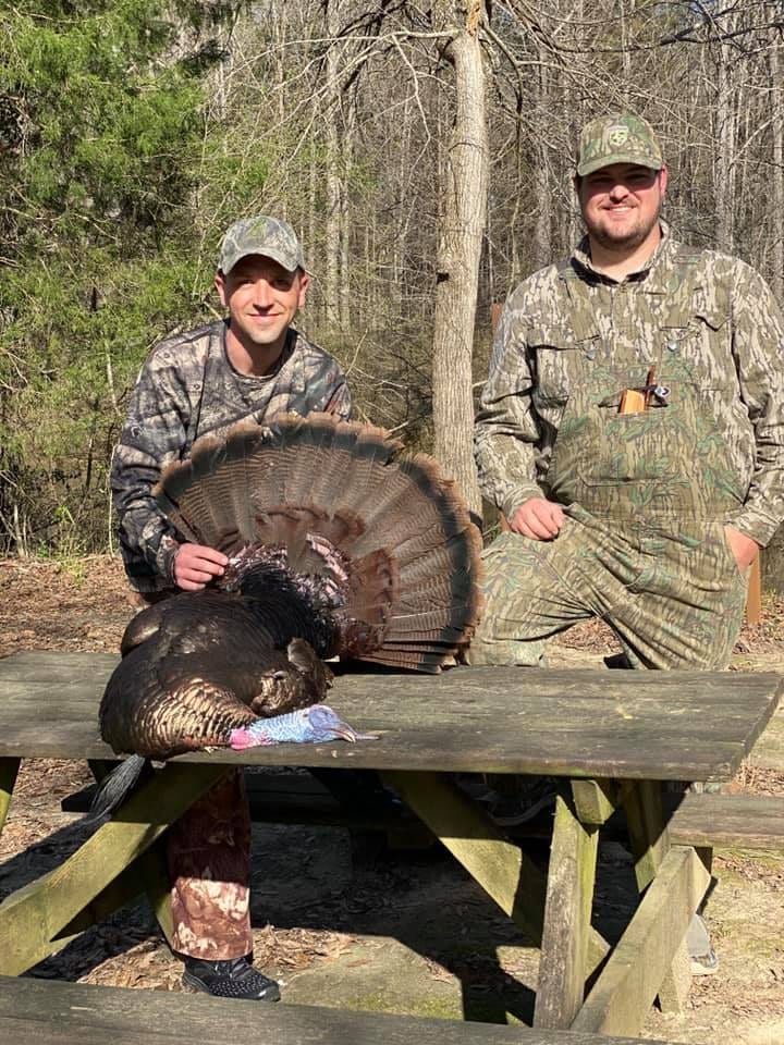 Two hunters pose with a harvested wild turkey on a picnic table.