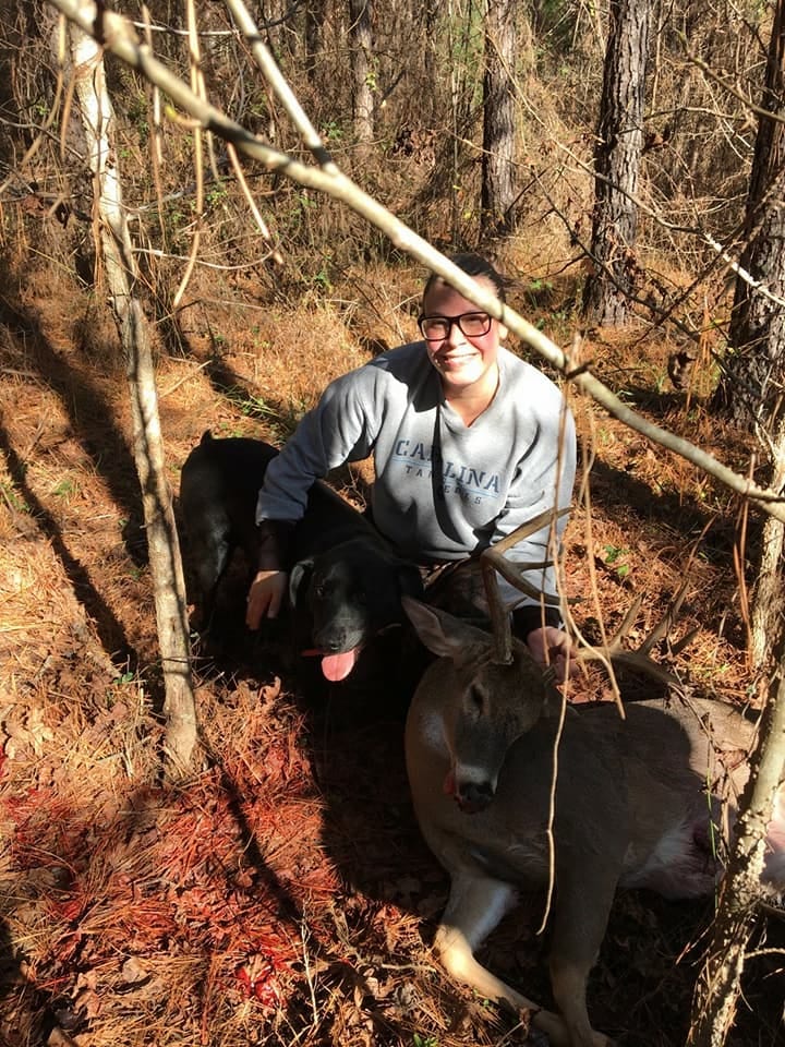 A hunter poses with a harvested whitetail deer and a dog in a wooded area.