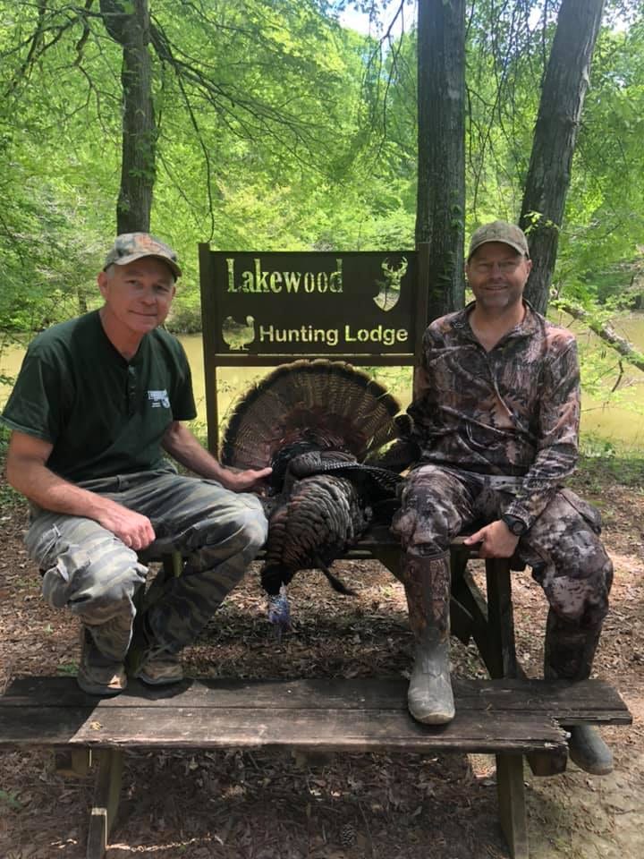 Two hunters pose with a harvested wild turkey in front of a Lakewood Hunting Lodge sign.