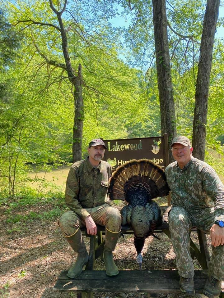 Two hunters pose with a harvested wild turkey in front of a Lakewood Hunting Lodge sign.