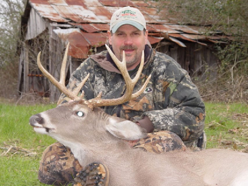 A hunter poses with a harvested whitetail deer in front of an old barn.