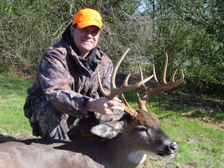 A hunter in camouflage and an orange hat poses with a harvested whitetail deer.