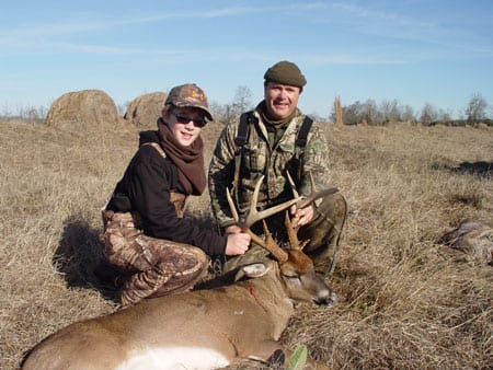 Two hunters pose with a harvested whitetail deer in a field.