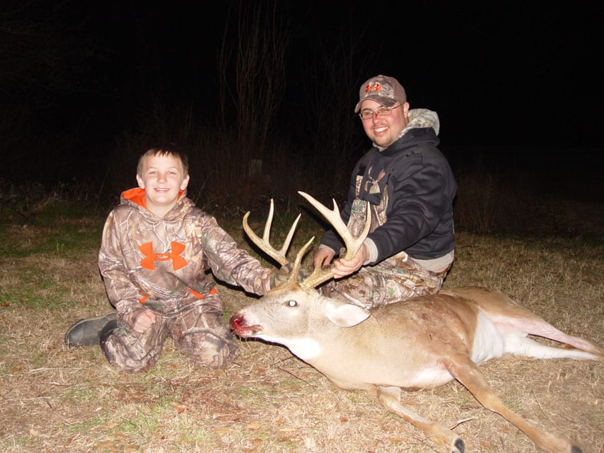 A man and a boy pose with a harvested whitetail deer at night.