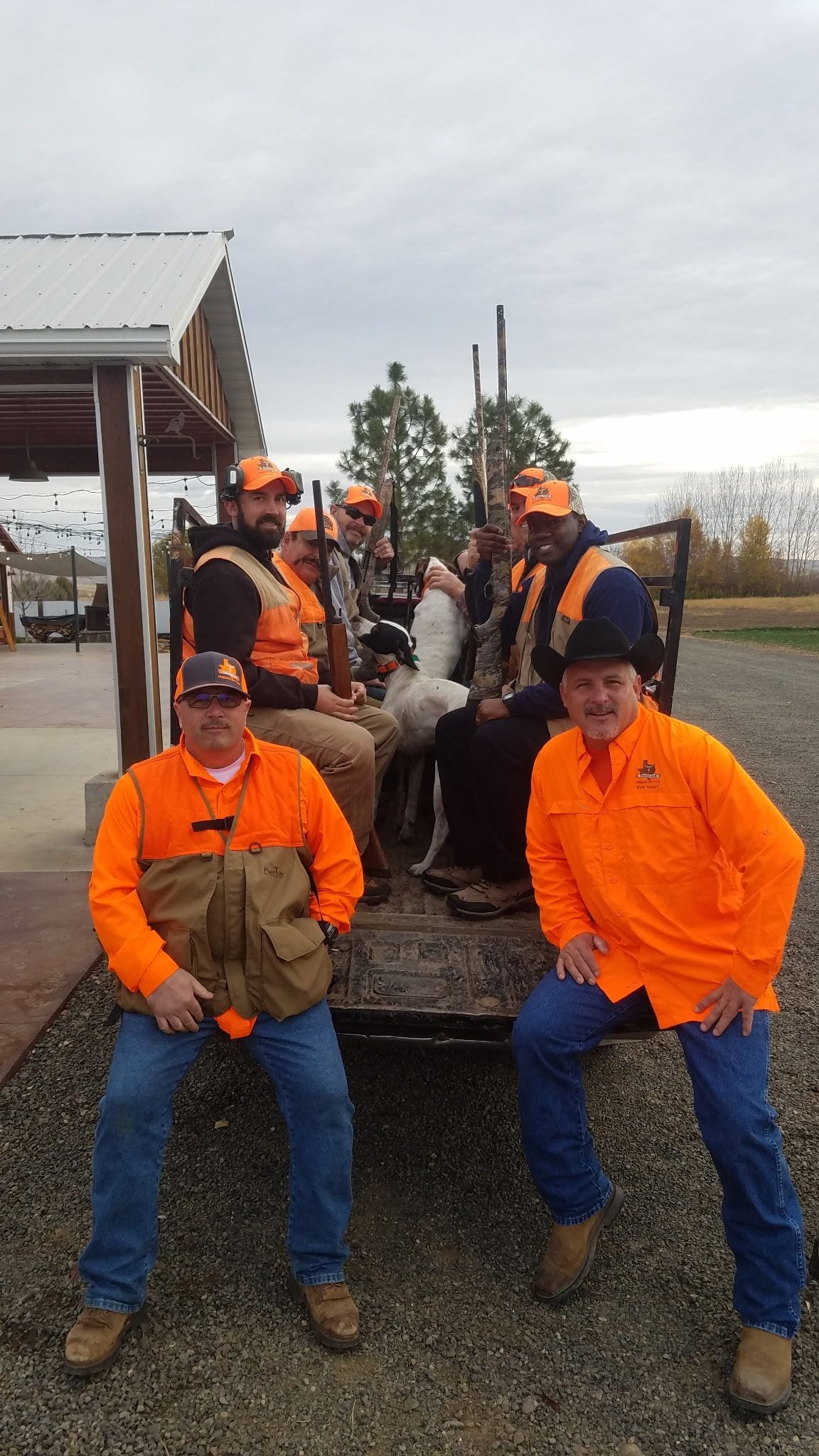 Two men in bright orange gear pose in front of a row of workers on a wagon outdoors.