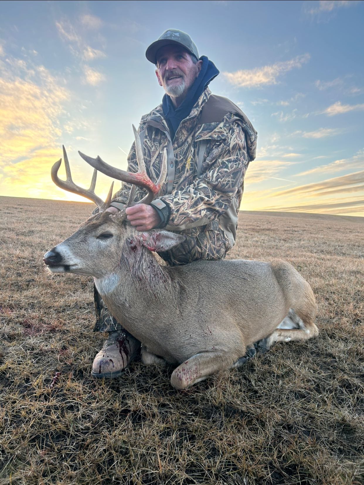 A hunter in camouflage poses with a harvested whitetail deer in a field at sunset.