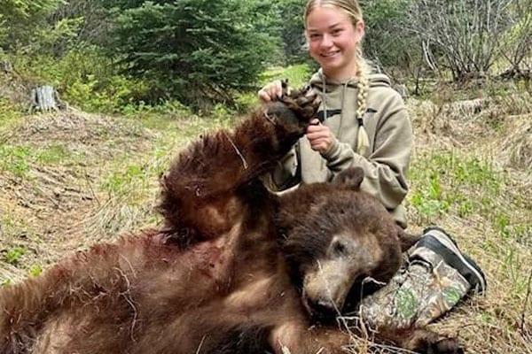 A young woman poses with a harvested bear in a grassy, wooded area.