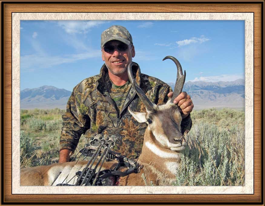 A hunter in camouflage clothing poses with a harvested pronghorn in a field with mountains in the background.