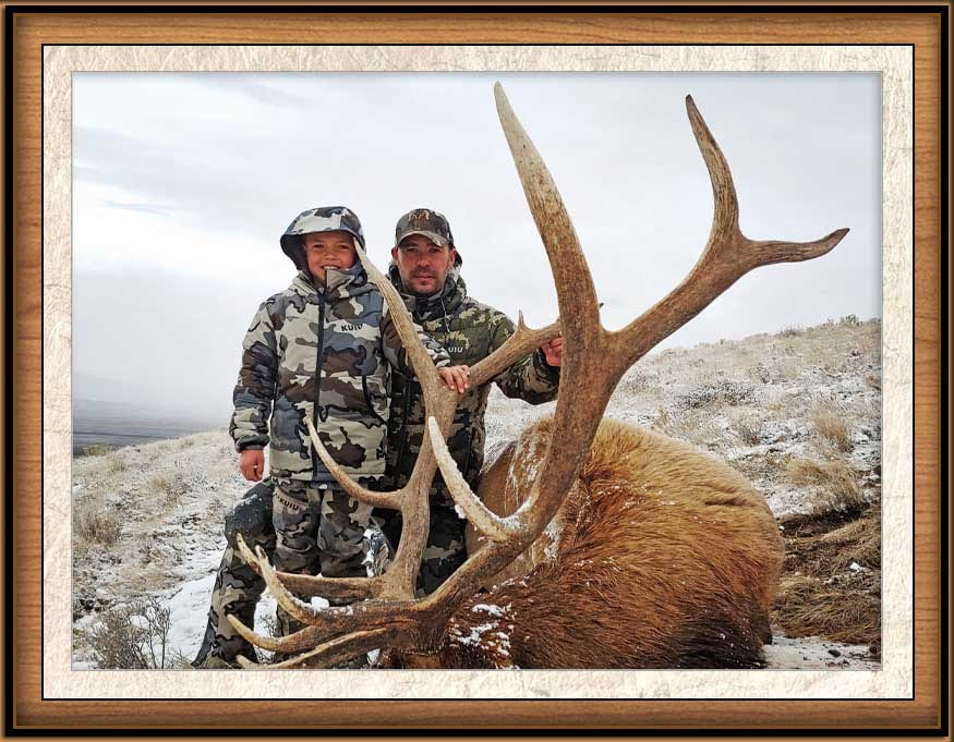 A hunter and a child pose with a harvested elk in a snowy field.