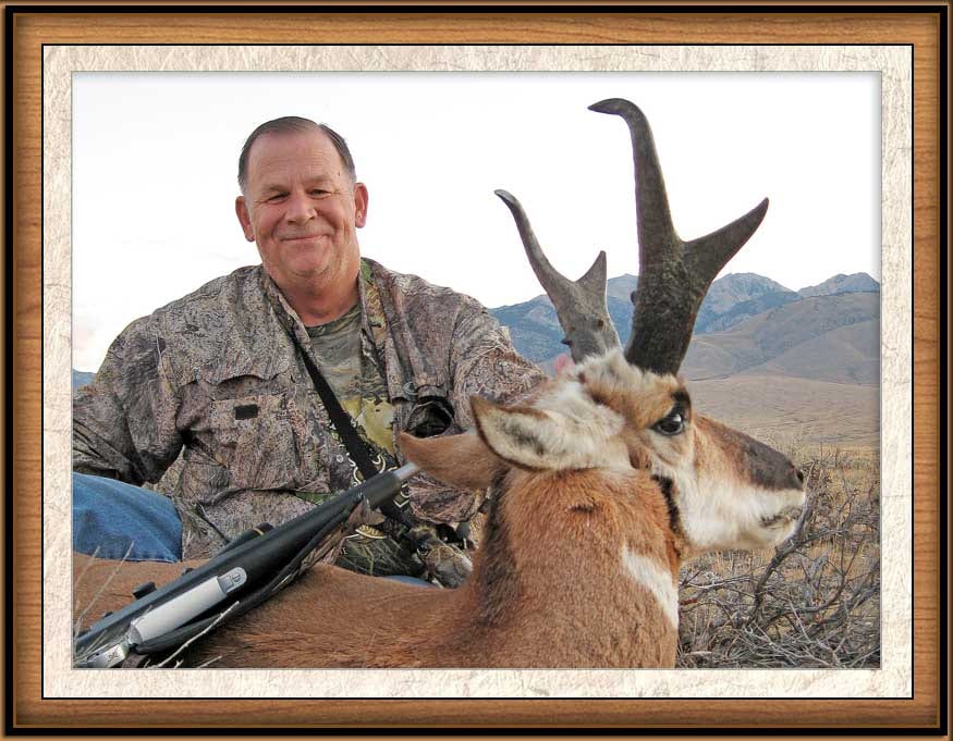 A hunter in camouflage poses with a harvested pronghorn in a field with mountains in the background.
