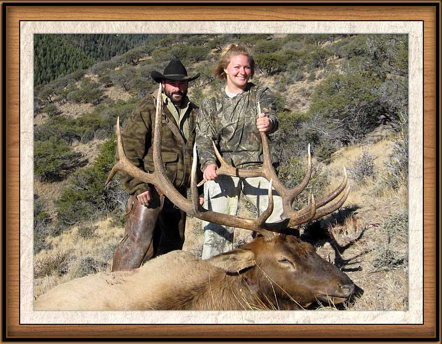 A man and a woman pose with a harvested elk in a mountainous, brushy landscape.