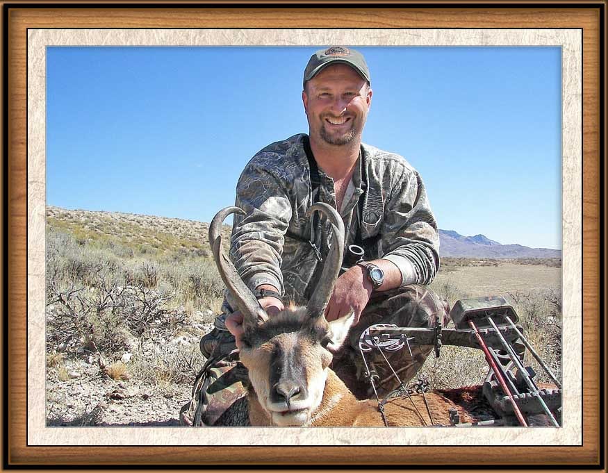 A hunter in camouflage clothing poses with a harvested pronghorn in a dry, open landscape.