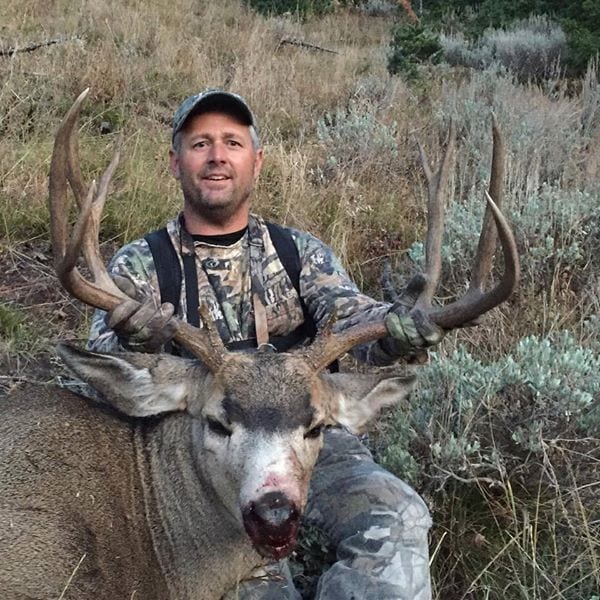 A hunter in camouflage poses with a harvested mule deer in a grassy, brushy field.
