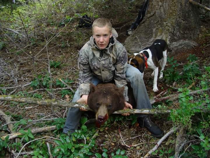 A young man in camouflage kneels with a harvested black bear, with a dog standing behind him in a wooded area.