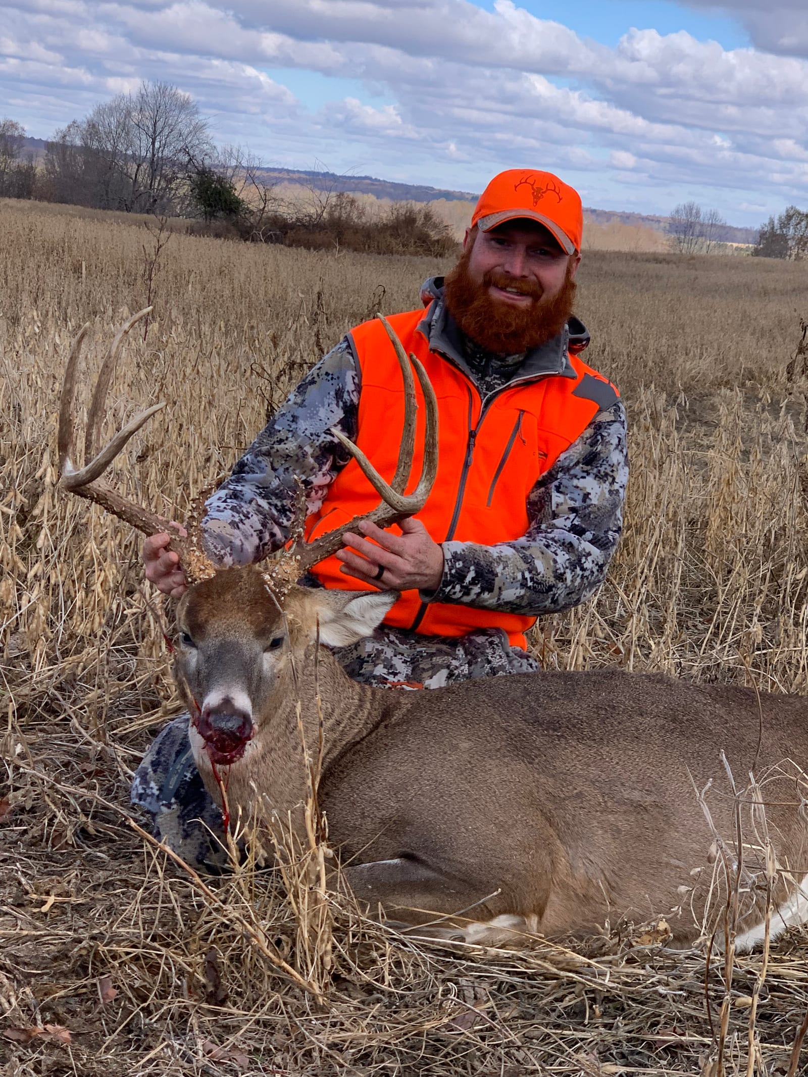 A hunter in an orange vest and camouflage kneels in a field, posing with a harvested whitetail deer.