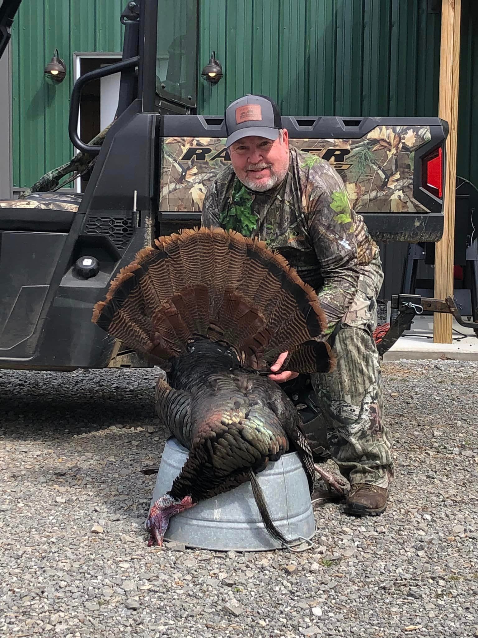 A man in camouflage holds a harvested wild turkey next to a UTV.