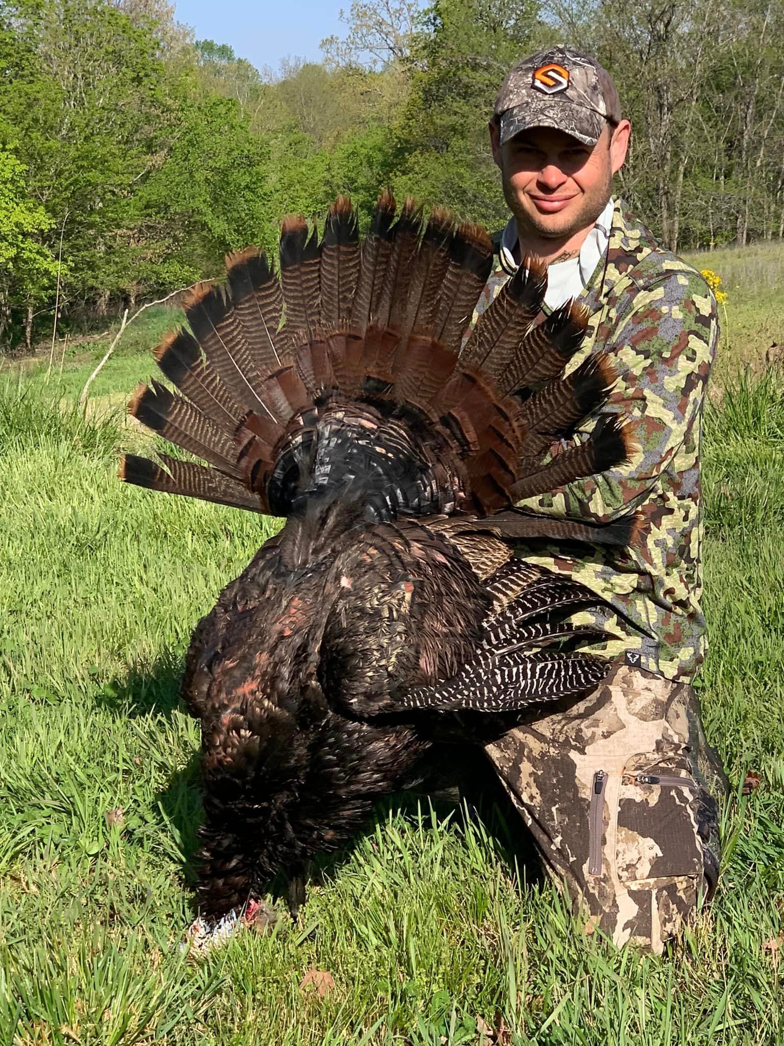 A hunter kneels in a grassy field, holding a harvested wild turkey with its tail fanned out.