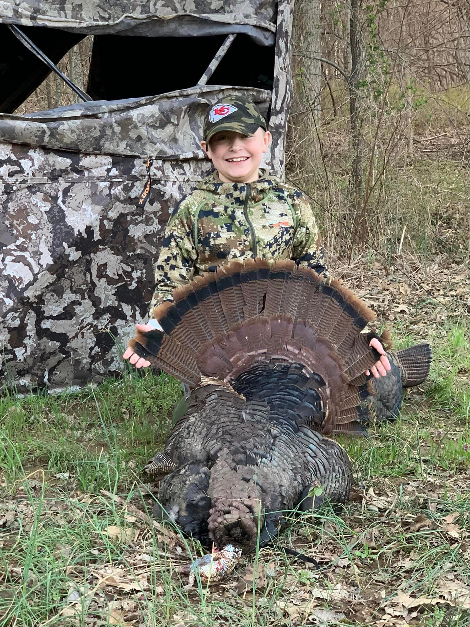 A young boy in camouflage poses with a harvested wild turkey in front of a hunting blind.