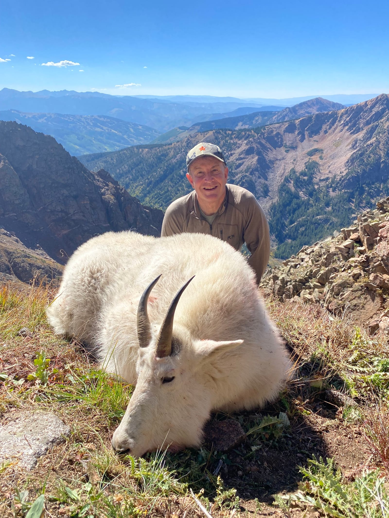 A man in outdoor gear smiling behind a dead mountain goat with curved horns, lying on the ground amid grassy terrain in a mountainous landscape.