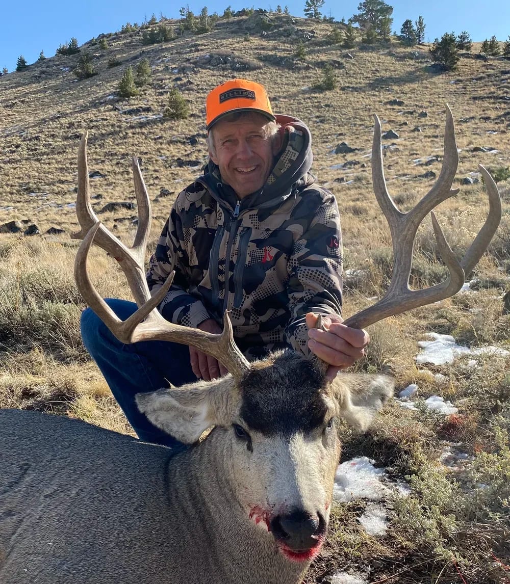 A man outdoors in a mountainous area holding the antlers of a freshly hunted elk with a laceration on its nose, wearing camouflage jacket and orange cap, smiling at the camera.