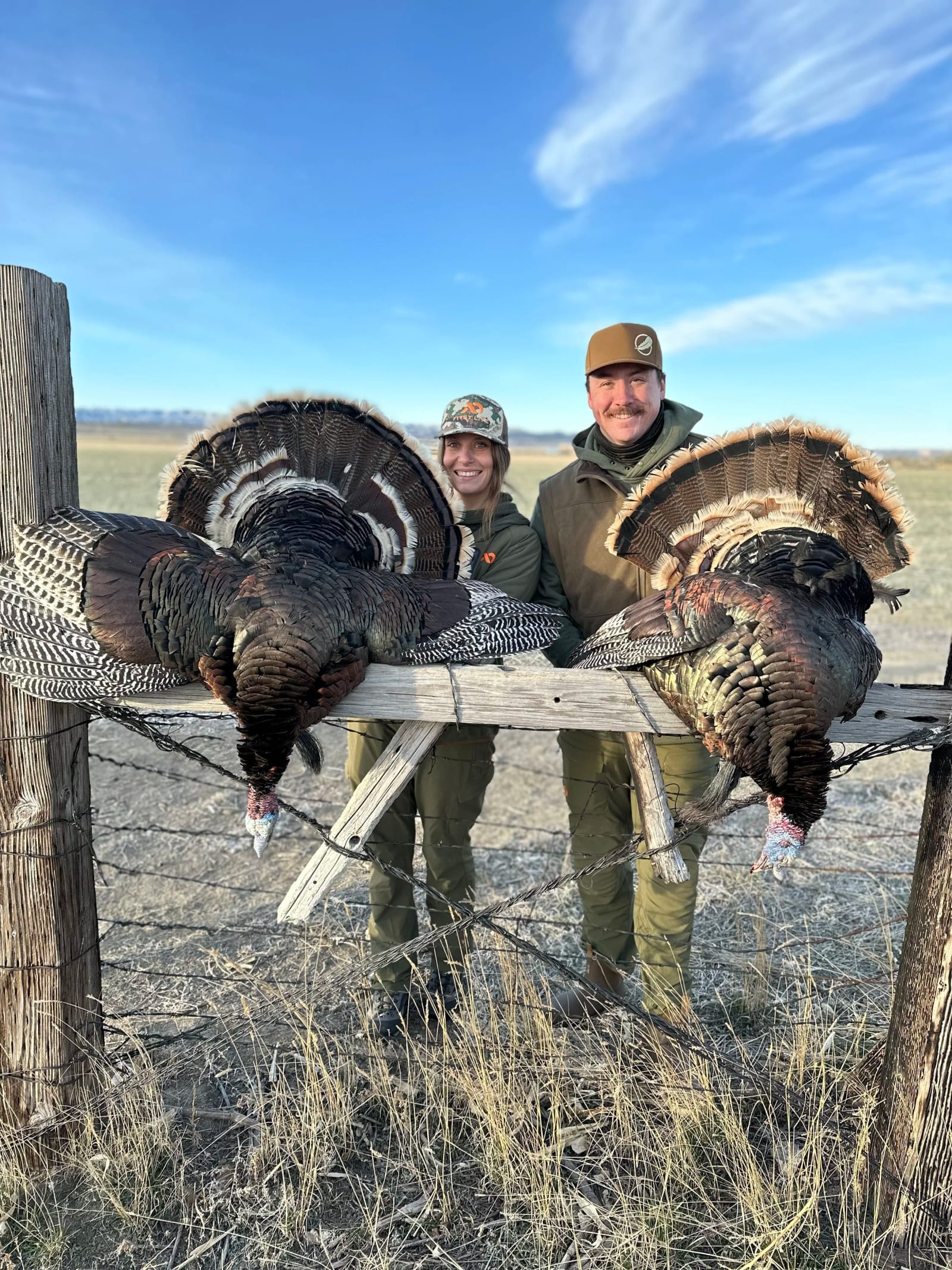 Two hunters, a man and a woman, smiling and posing behind two large turkeys they have hunted, outdoors in a rural area.