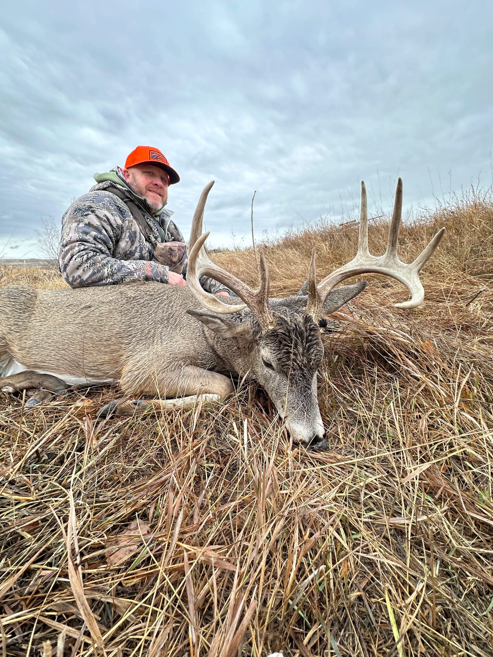 A man in camouflage clothing and an orange cap kneels next to a large, dead elk with impressive antlers, lying on dry grass under a cloudy sky.