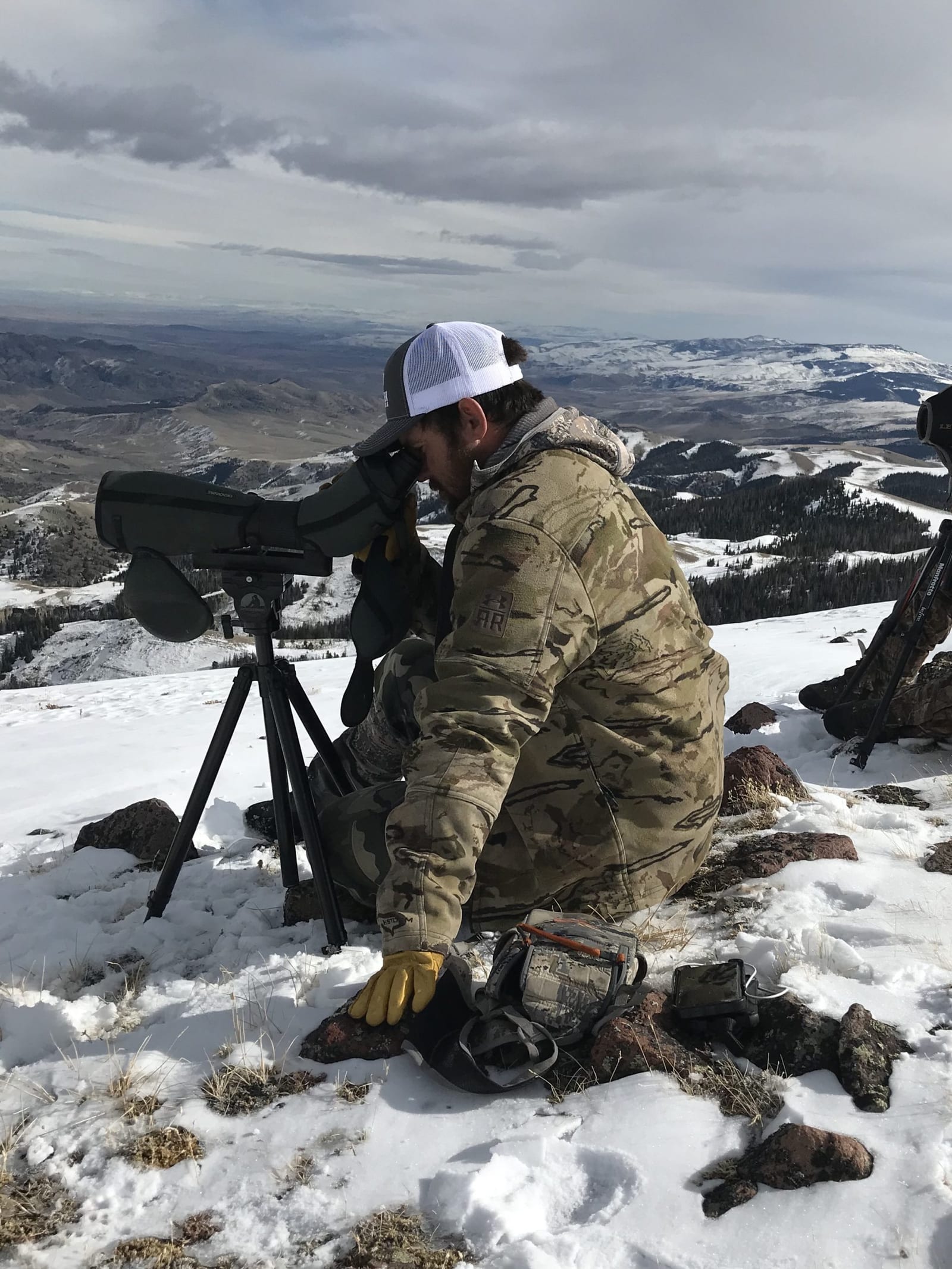 A hunter in camouflage looks through a spotting scope on a snowy mountain, with harvested animal parts visible on the ground.