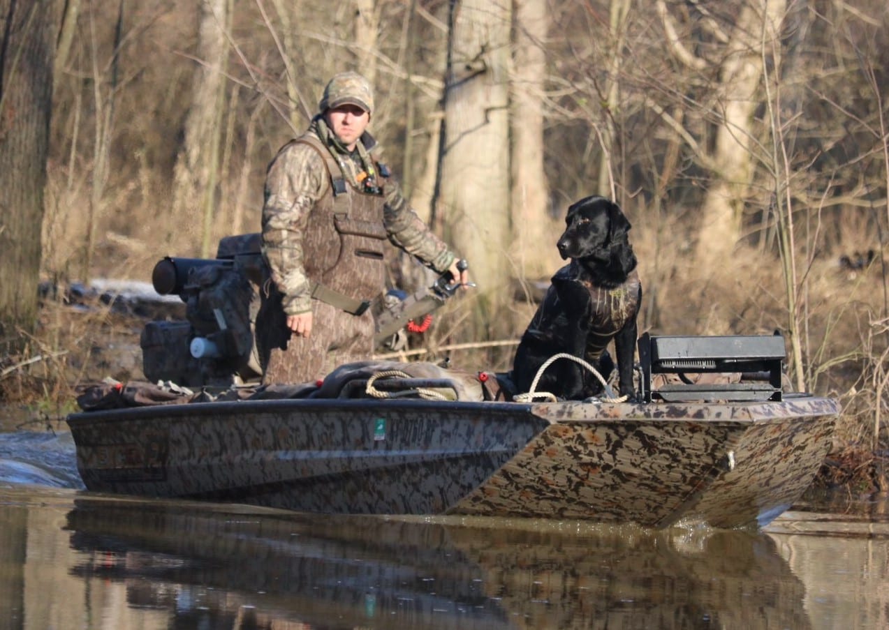 A hunter and a black dog are in a camouflage boat on the water, with trees in the background.