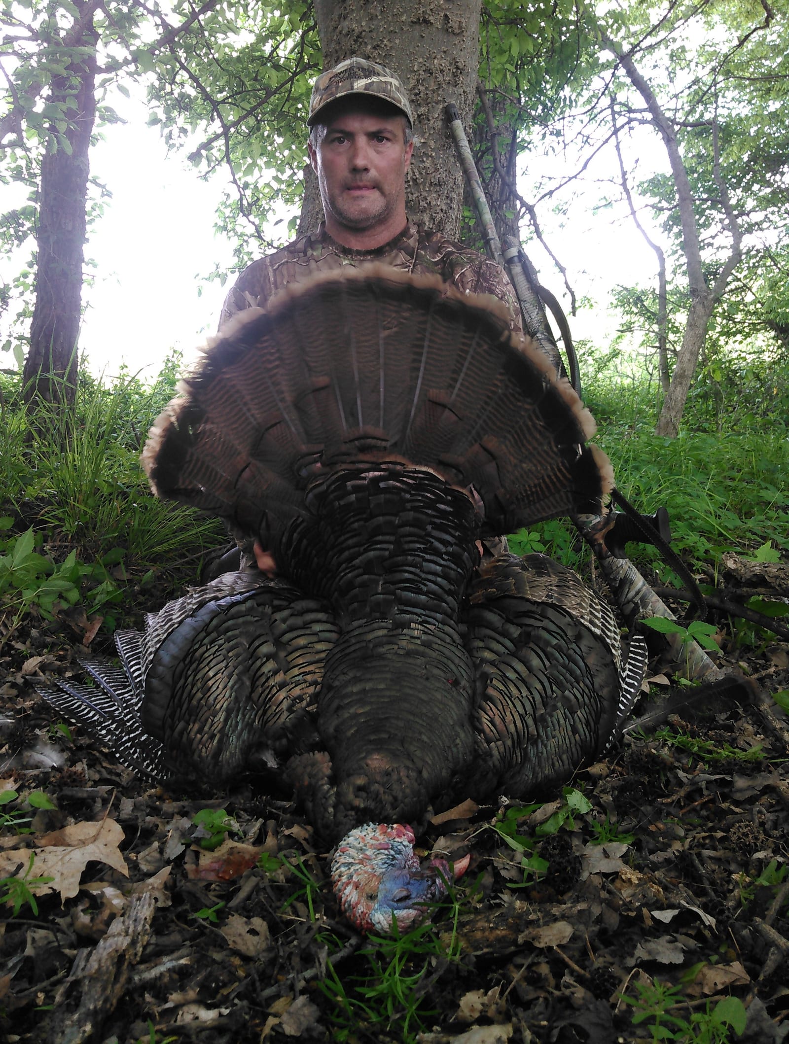 A hunter poses with a harvested wild turkey in a wooded area.