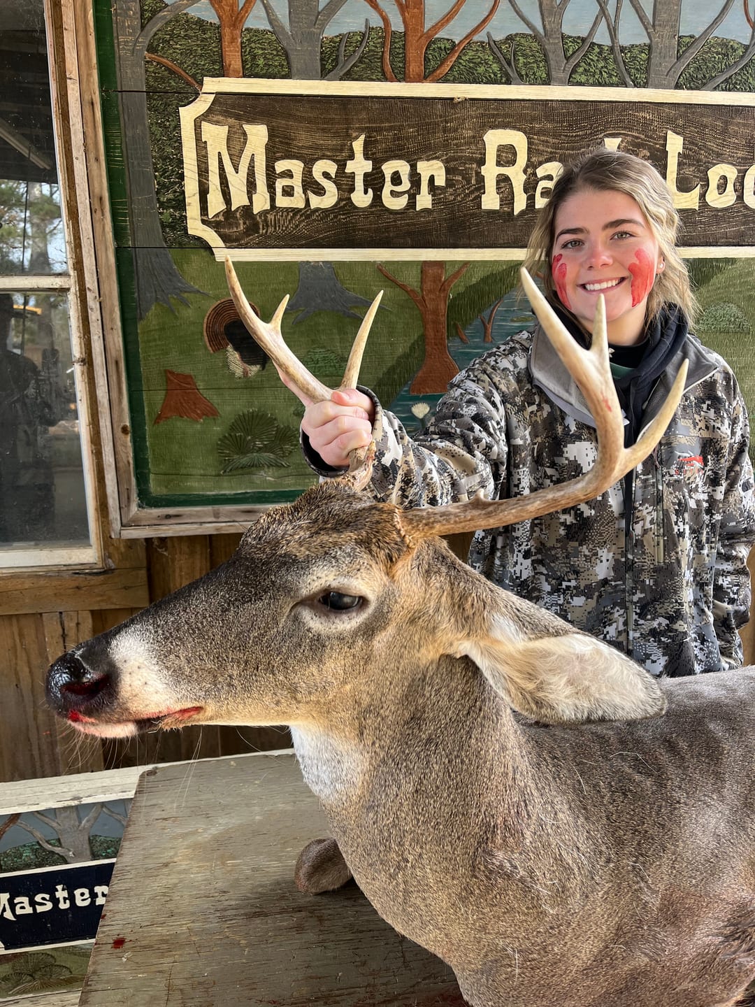 A young woman poses with a harvested whitetail deer in front of a Master Rack Lodge sign.